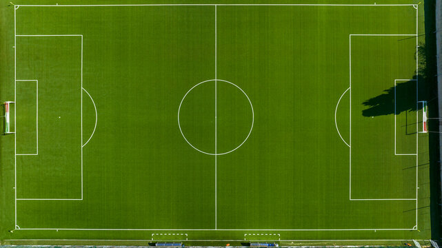 Perpendicular top-down aerial view of a vibrant green synthetic soccer field, showcasing precise white boundary lines, the center circle, penalty areas, and goals under clear, bright daylight.