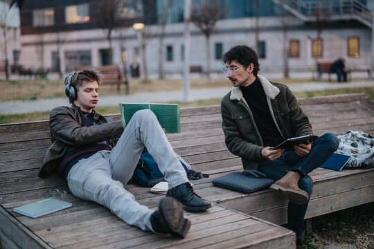 Two high school students sit on a park bench studying with notebooks, headphones and a tablet. The friends review notes together outdoors while one listens to music and the other uses a tablet.