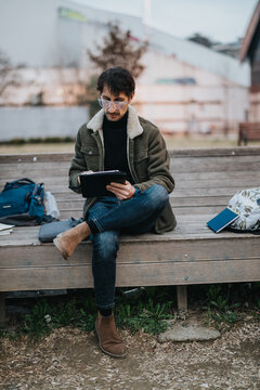 A high school student sits on a bench studying on a tablet while wearing glasses and a jacket. The young man concentrates on digital homework outdoors in an urban park setting.
