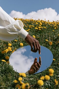 Hand reaching toward mirror on grassy field with dandelions