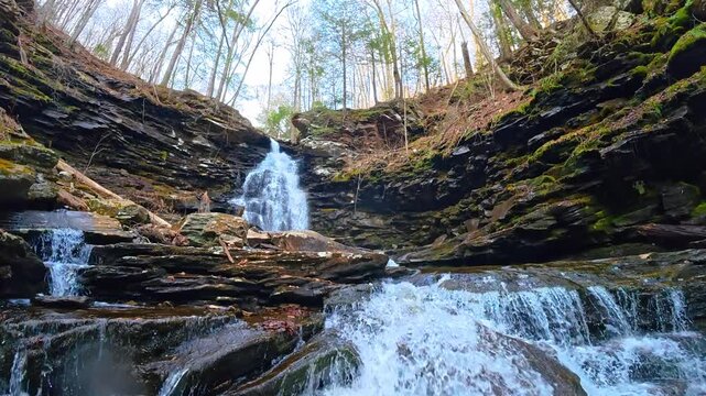 Wide view of multi tiered waterfall cascading down rocks. Scenic forest waterfall falling over dark stone ledges into a stream surrounded by trees.