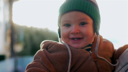 Baby smiling in bear teddy fleece coat and green beanie hat while held outdoors, joyful infant enjoying winter day in parent’s arms with warm family connection and happiness © Marco