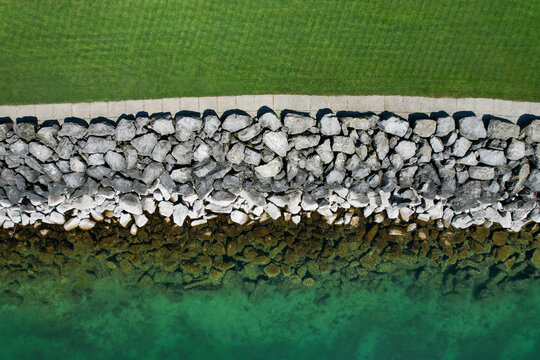 Aerial view of the rocky shoreline of Lake Geneva featuring a stone breakwater, a paved walkway, and lush green grass Morges, Vaud, Switzerland.