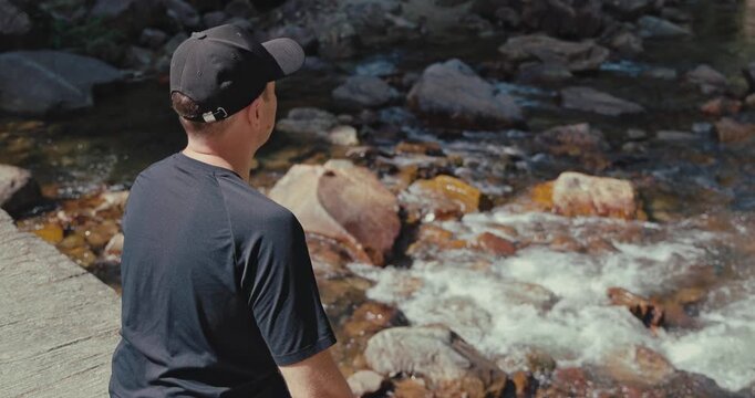 Man wearing black cap sits on stone bridge looking at flowing river water. Outdoor adventure and relaxation in nature during summer hike. Back view of male traveler.