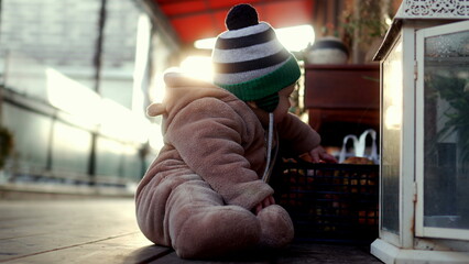 Baby sitting in bear teddy fleece coat and striped beanie hat outdoors playing near crate of apples, toddler exploring surroundings with curiosity during cozy cold season © Marco