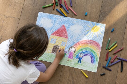 Young child drawing colorful family house scene with rainbow and smiling cartoon characters using pastel chalks on large paper, creative kids activity, cheerful indoor setting with scattered art