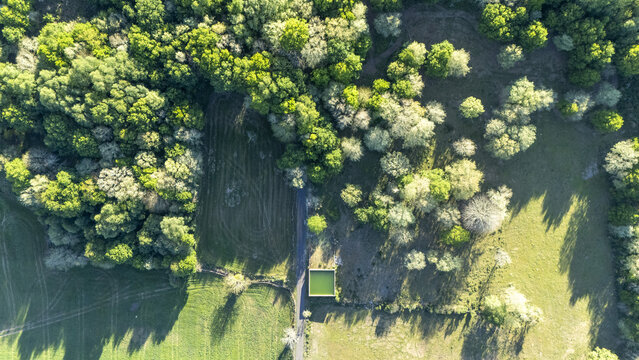 Aerial view of lush green forest canopy, open meadows, and a small square water tank beside a narrow rural road Ourense, Province of Ourense, Spain.