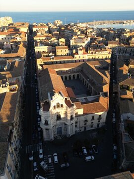 Aerial view of the historic Church of San Placido and its courtyard surrounded by traditional rooftops and a long street leading to the harbor under golden hour light in Catania, Sicily, Italy.