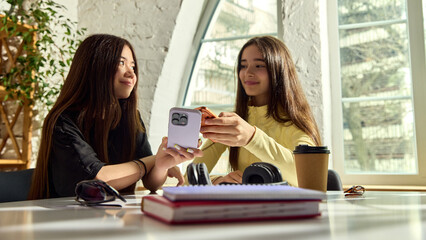 Fototapeta na wymiar Teenage girls sharing smartphones for money transfer near notebooks. Concept of digital allowance, peer payment, student budgeting, online checkout and safe cashless habits.