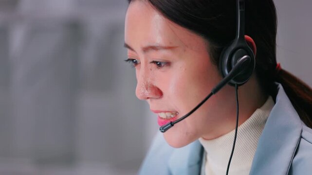 Friendly Asian female call center agent providing online customer support while typing on laptop in modern office environment. Professional customer service and communication concept.
