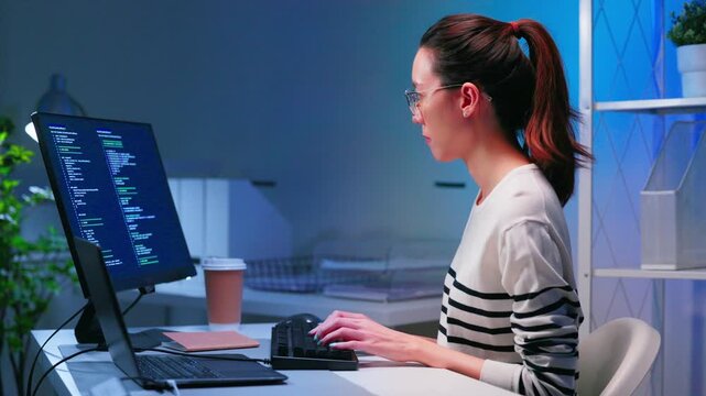 Focused Asian woman coding on computer during overtime night shift in modern office. Software engineering, startup workflow, remote development and women in technology concept.
