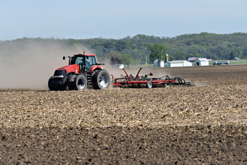 Fototapeta premium Red tractor Pulling a Double Fold Cultivator for spring field preparation with a farm scene in the background.