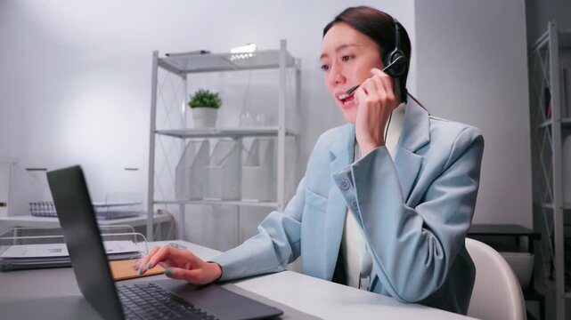 Friendly Asian female call center agent providing online customer support while typing on laptop in modern office environment. Professional customer service and communication concept.