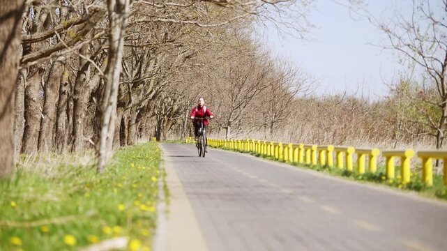 Young adult woman with red jacket cycling on rural road lined with bare trees under bright daylight
