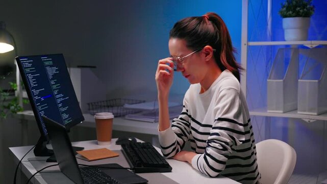 Focused Asian female programmer working overtime at night while coding on computer in modern office. Software development, deadline pressure, startup culture and women in tech concept.