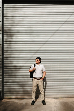 Man Standing Against Corrugated Metal Door, Carrying Messenger Bag, Wearing Sunglasses And White Shirt, Khaki