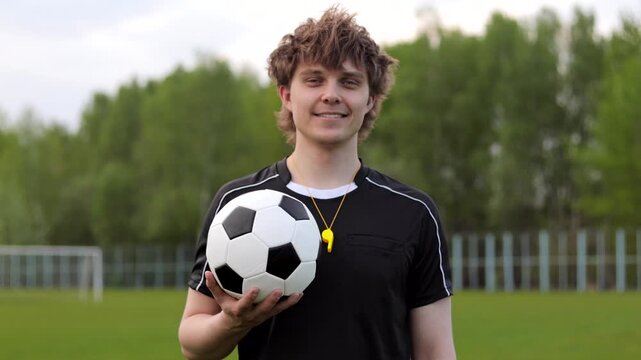 Young smiling soccer referee or coach holding a football in black referee uniform. Concept of officiating, fair play, sports rules and game control.