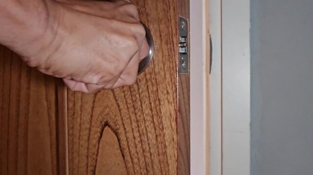 A hand reaches out to turn a round stainless steel doorknob on a modern light-brown wooden door