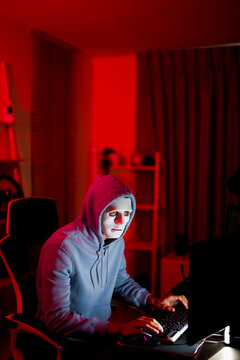 Anonymous crypto hacker in white mask using computer in red neon room, cyber security threat and online hacking concept, masked person typing on keyboard with dramatic lighting and digital crime