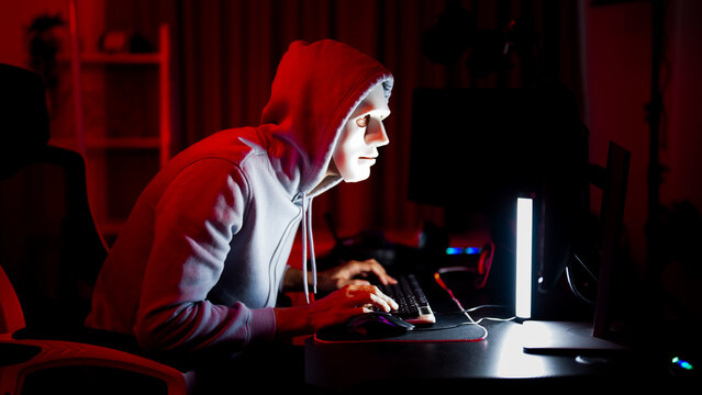 Romance scam wearing white mask and hoodie using computer in dark red room, cybercrime, cybersecurity threat concept, masked person typing on keyboard with dramatic neon lighting and online attack