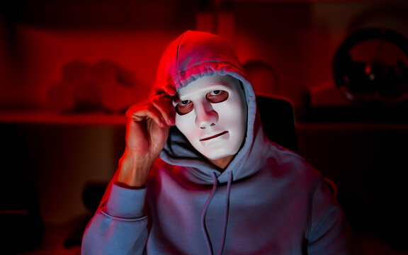 Anonymous hacker wearing white mask and hoodie in dark red room, cyber security threat and online crime concept, mysterious masked person with dramatic lighting, digital privacy and hacking danger
