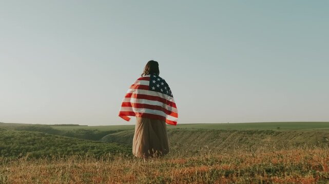 Woman Wrapped In American Flag Standing On Windswept Bluff Overlooking Green Valley At Golden Hour, Traveler