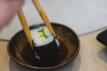 Close-up of wooden chopsticks dipping a cucumber sushi maki roll into soy sauce. Concept of Japanese cuisine, healthy eating, and traditional oriental dining. Action shot of eating sushi in a restaura © Anna Kondratiuk