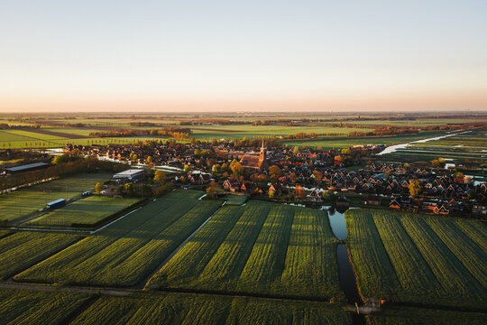 Aerial view of the historic village of Schermerhorn with its church spire surrounded by lush green polder fields and narrow canals during golden hour in Schermerhorn, North Holland, Netherlands.