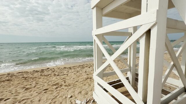 A white lifeguard structure frames a beautiful view of the sandy shore and foaming waves. The scene feels open and refreshing, offering a sense of calm protection by the vast, breezy ocean.