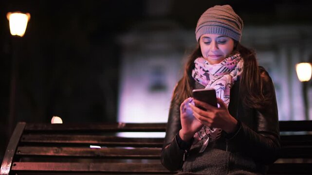 Young Woman Sitting On Bench Using Smartphone At Night With Warm Lights Around Representing Mobile Communication Relaxed Lifestyle And Digital Interaction In Urban Environment