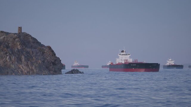 Camera pans along stony Headland to reveal Crude Oil Tanker Ships anchored in Bay. Aerial View of Oil Tankers anchored in the Ocean at Sunset. Strait of Hormuz Blockade Concept. 3d Animation Render