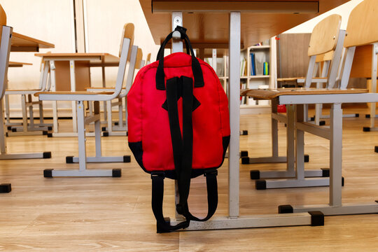 Red schoolbag hanging on wooden desk in empty classroom. Backpack on table in school class interior. Back to school concept, elementary education, student supplies, learning furniture, copy space