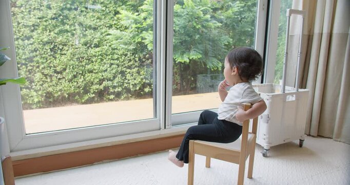 A curious toddler sits alone on a chair, thoughtfully looking out the window at the garden, experiencing a moment of quiet observation and early development.