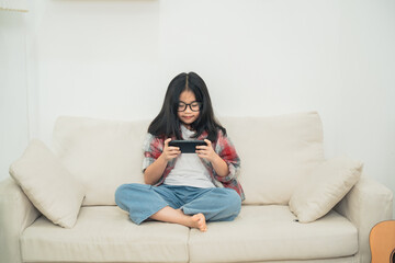 Fototapeta premium Young girl with glasses sitting on a couch playing a mobile game, wearing casual clothes, enjoying leisure time indoors with a relaxed atmosphere