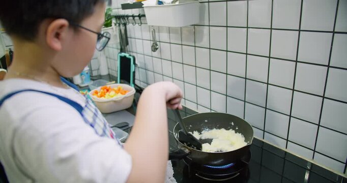 A young boy learning to cook stirs ingredients in a frying pan on a modern gas stove, developing practical life skills through a hands-on home cooking lesson.