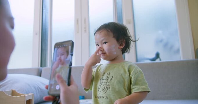 An adorable toddler with face paint playfully poses for a smartphone camera as a parent records a video for social media content creation in a modern home.