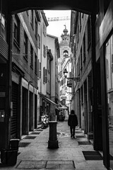 Naklejka premium Pedestrian walking in a narrow alley toward a bell tower in a black and white urban scene.