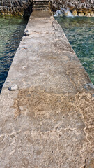 Fototapeta premium A narrow aged concrete path stretches across turquoise sea water leading to small stone stairs and a wall on a sunny day