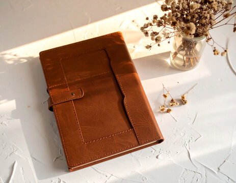 A closed brown journal rests on a textured white surface beside a vase with dried flowers