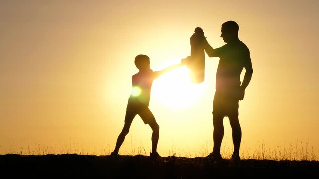 Father and son silhouette at sunset. Dad and child share joyful moments with a beach towel. Family bonding creates warm atmosphere. Outdoor activity promotes happiness and connection.