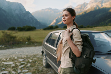 Obraz premium Smiling woman with backpack beside a car in a valley with mountains in the background, wearing a casual jacket, enjoying an outdoors lifestyle, travel and adventure vibe under natural light.