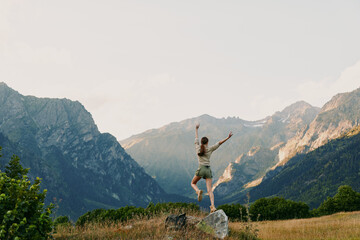 Obraz premium Young woman stands on a rocky outcrop with arms raised in celebration, framed by sunlit alpine valley and distant mountains, conveying freedom and a sense of adventure.