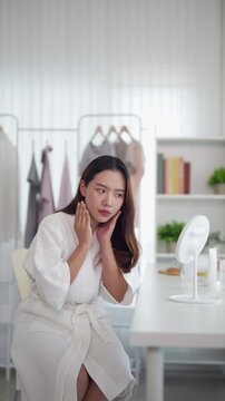Asian women beauty routine skincare applying facial massage touching face morning ritual sitting white bathrobe home bedroom vanity mirror background clothing