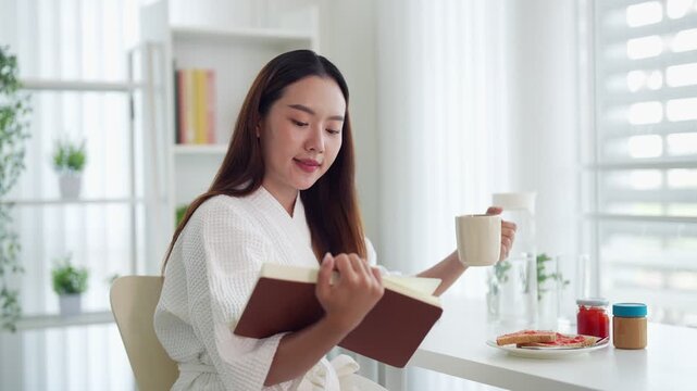 Asian women reading book drinking coffee relaxing at breakfast morning routine wearing white robe sitting in bright modern kitchen apartment enjoying leisure