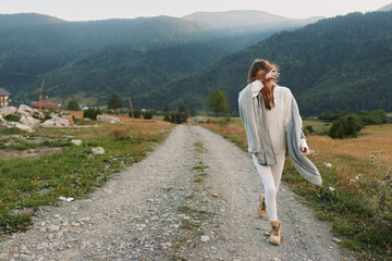 Naklejka premium Woman outdoors, lifestyle scene on a mountain path, wearing a cardigan, white pants, boots, smiling with relaxed posture, sunlit breeze, natural colors, vast scenery