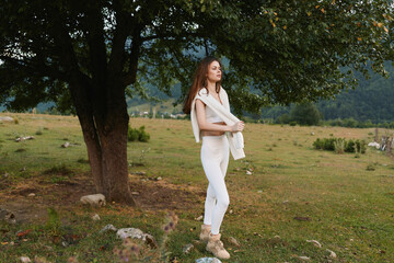 Naklejka premium Young woman in white outfit standing near a tree in a green meadow. Outdoor lifestyle scene with nature and calm mood during daytime.