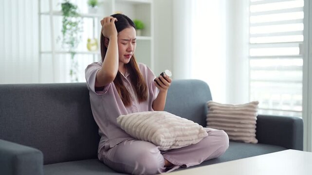 young Asian women stressed about hair loss holding smartphone sitting on sofa at home living room checking scalp health problem concern worry about beauty
