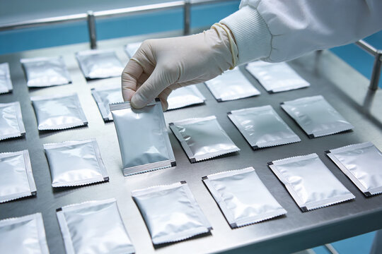 Gloved hand selects sealed silver foil packets from a tray in a clean room