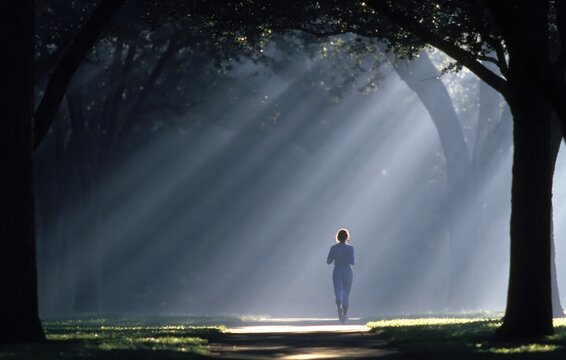 Woman jogging in sun rays through foggy forest path