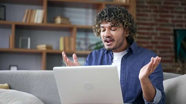 Angry excited young man arguing on video call using laptop computer sitting on sofa in living room at home. Dissatisfied stressed male emotionally talks shouts about problems expressing anger Close up